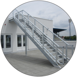 A silver metal staircase rises diagonally along the side of a light-colored building. A landing and guardrails connect the upper level to the paved ground. Glass doors and windows line the lower facade. A cloudy sky is visible in the background.