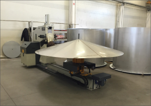 A broad conical sheet of metal rests on machinery in a workshop. A large cylindrical metal tank stands immediately behind it on the right. Additional equipment and a small metal coil are visible around the work area. The floor is pale gray concrete.