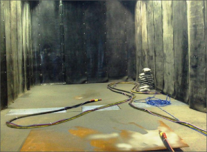 The image shows the interior of a tall enclosed work area with dark, stained wall panels. Several hoses or cables lie across the concrete floor and curve toward the rear. A small striped object is visible near the back-right wall. Light enters from the upper left.