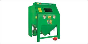 A green box-shaped machine stands on legs against a white background. Its slanted upper face has two circular viewing windows. A small control panel sits near the top edge. The lower section narrows into a funnel above a small red component.