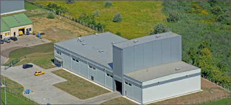 An elevated oblique view shows a large industrial building with several joined gray roof sections. A paved yard and small parked vehicles sit beside the building. Grass, trees, and fenced land surround the site.