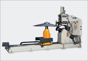 A gray industrial machine is shown against a white background. A long narrow rail extends left from its central assembly. An orange cylindrical component rises beneath a circular metal plate. An upright control unit with visible cables stands on the right.