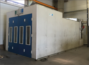 A large rectangular industrial enclosure fills much of a workshop. Its blue front panel has several tall, narrow windows. White side panels extend toward the back. Overhead beams and other workshop equipment are visible around it.