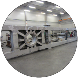 A large rectangular metal frame with a circular opening at one end sits across a workshop floor. Long metal rails and equipment extend behind it to the right. Bright ceiling lights illuminate the work area. The image is framed within a circular crop.
