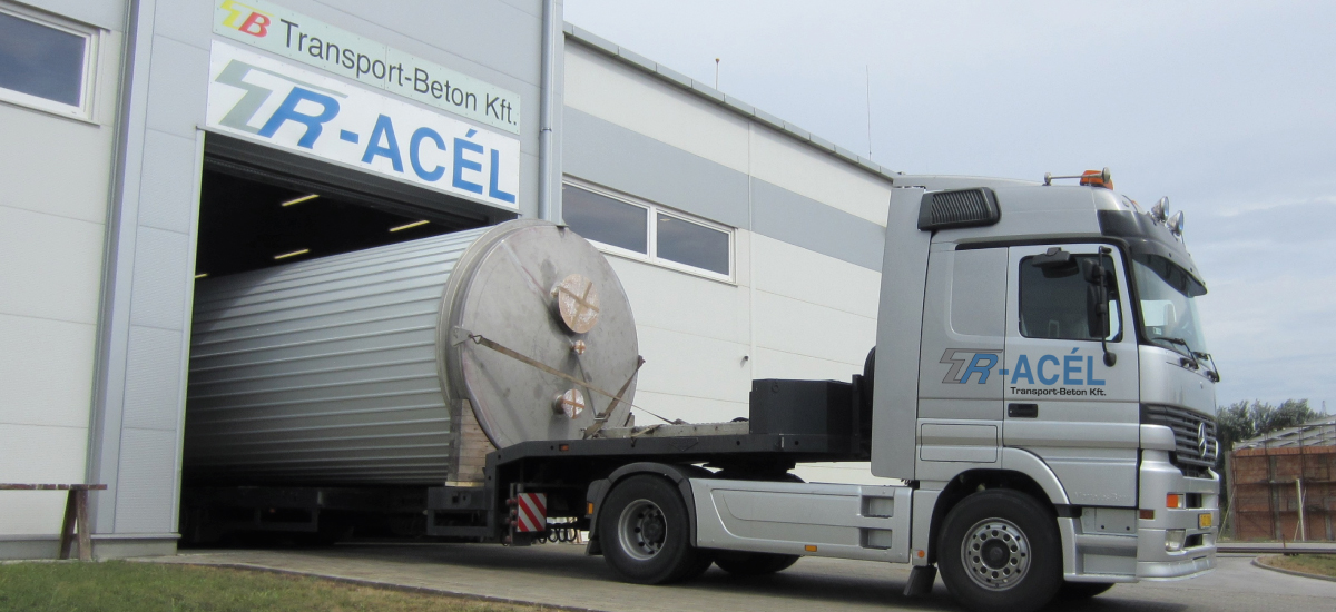 A silver tractor truck stands outside a wide industrial loading bay. A large cylindrical load rests on a trailer extending from the bay, with part of the load still inside the doorway. The building sign has Transport-Beton Kft. text, while TR-ACÉL appears on the truck door.