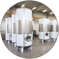 Several tall stainless-steel cylindrical tanks stand inside a bright workshop. The nearest tank has a round opening in its lower front and small fittings at the top. More tanks and overhead lights fill the background. The image is framed within a circular crop.