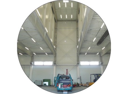 A tall workshop interior is framed by a large circular crop. High metal walls and the roof structure rise above a vehicle parked at the lower center. Bright strip lights and high windows illuminate the interior.